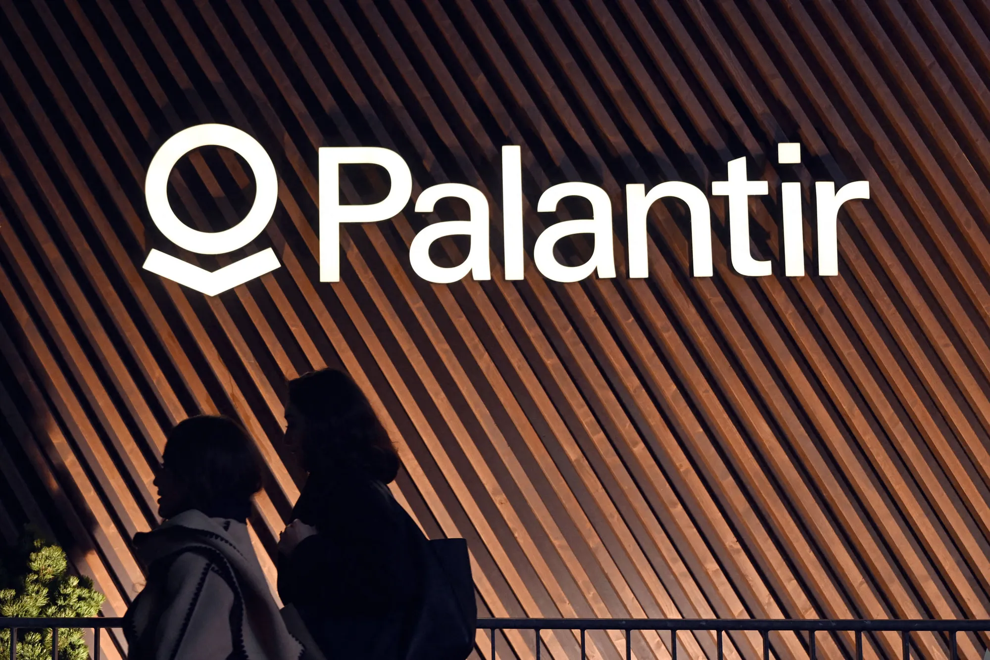 Two silhouetted pedestrians walking past an illuminated Palantir sign