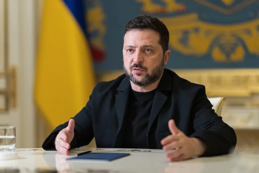 Ukrainian President Volodymyr Zelensky speaking at a table with the Ukrainian flag behind him